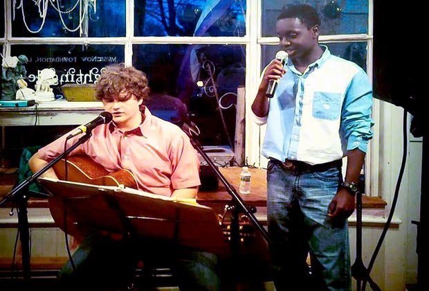 Image of one young man with a guitar and another with a microphone.